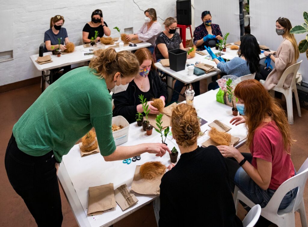 A group of workshop participants sit a tables in a recent kokedama workshop. They have small plants and coconut fibre in front of them. In the foreground Farm team member Kathleen leans over to help a particpant. 